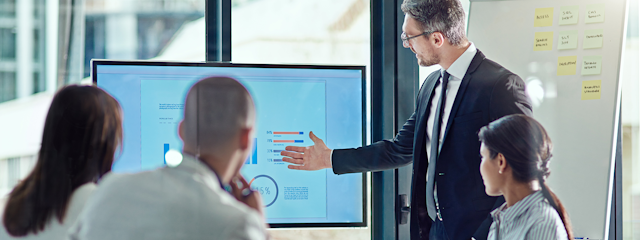 A person in professional attire gestures at charts on a screen while making a presentation.