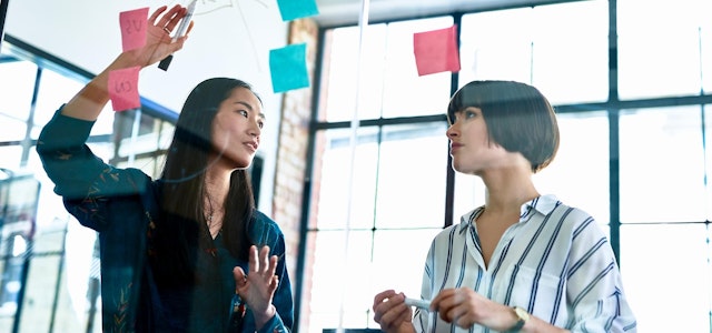 Two master's degree students working on a project with colorful sticky notes on a glass wall.