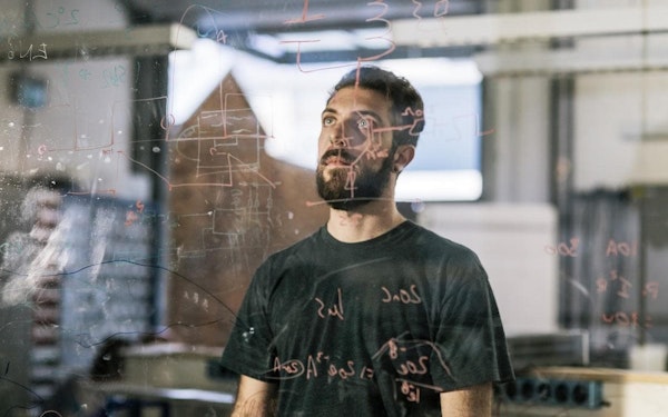 A man in a tee shirt stands behind a clear glass wall covered with equations and diagrams in marker.