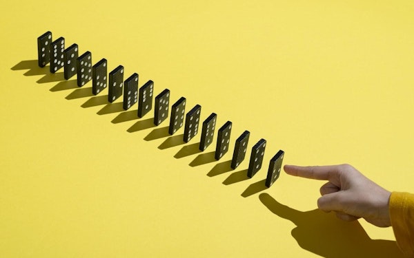 Against a yellow background, a hand is about to push a row of upright dominoes.