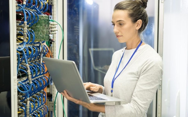 A computer network engineer works on their laptop in a server room.