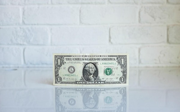 A single one-dollar bill stands upward against a white brick wall.