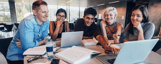 Graduate students smiling and working together at a table with laptops and open textbooks