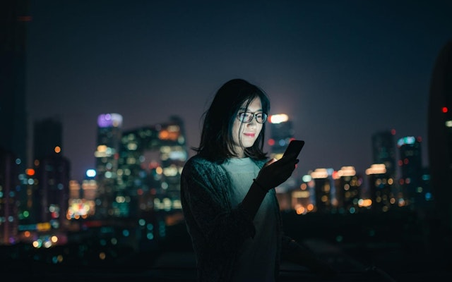 A young woman stands in front of a city skyline while looking at her phone.