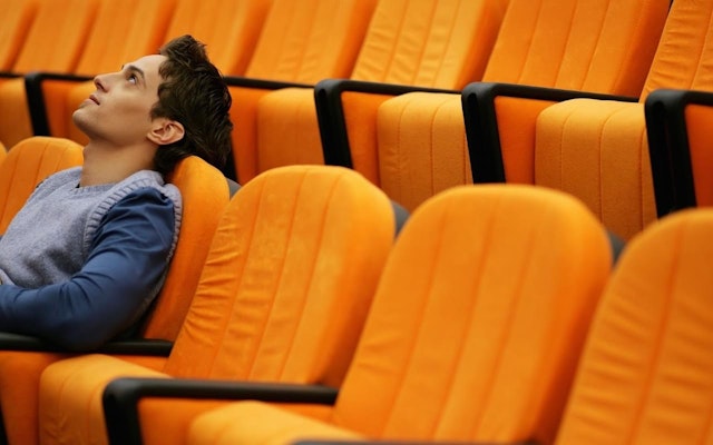 A male college student sits in a lecture hall with orange chairs while looking up.