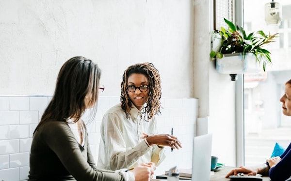 Three women sit and talk at a table with an open laptop.