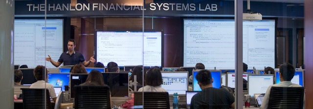Students in class at computers as the professor displays code on large screens.
