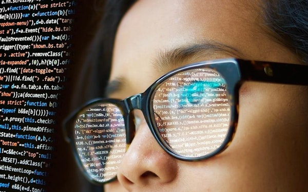 A screen with code is reflected in the glasses of a young person.
