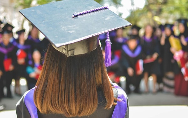 A student wears a black and purple graduation gown and cap while facing a crowd of peers at a graduation ceremony. 